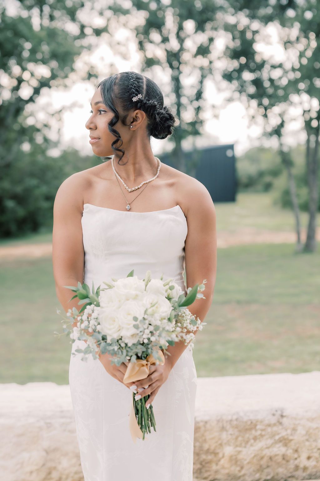 Elegant bride in strapless white gown holding white floral bouquet, gazing sideways outdoors amid lush greenery.