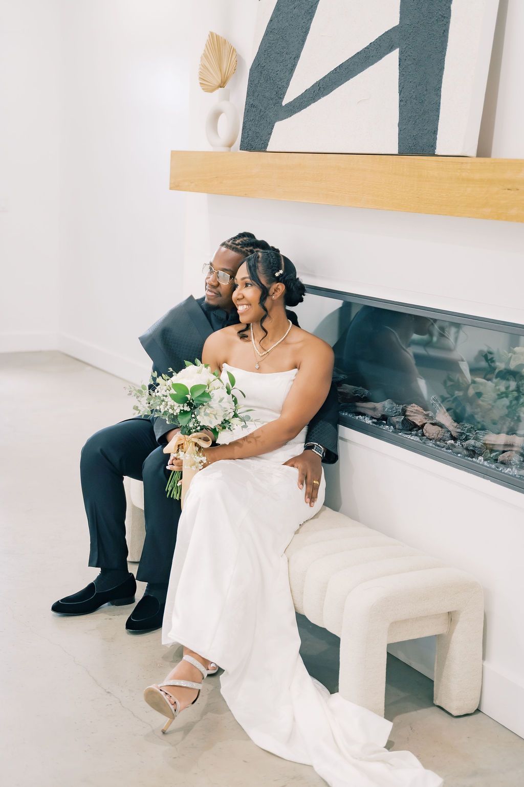 Bride and groom sit together by modern fireplace, bride holding white floral bouquet.