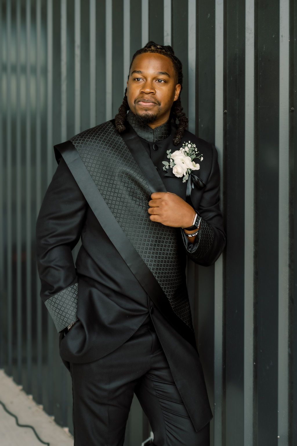 Man in black tuxedo with boutonniere leans against striped wall, wearing braids and formal attire.