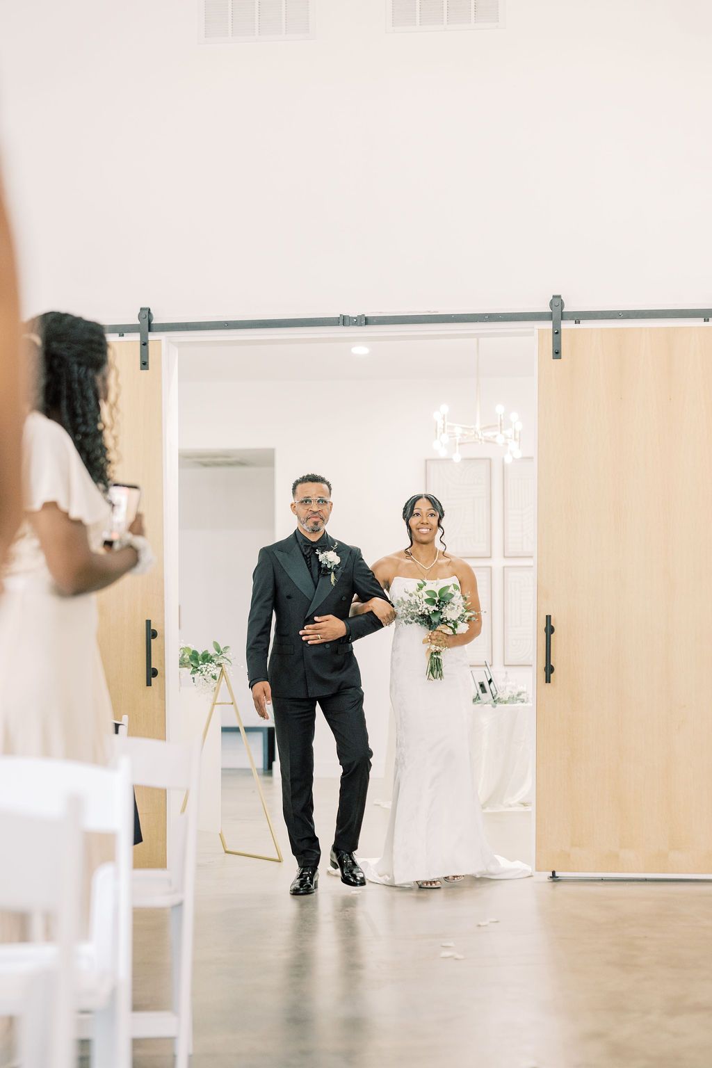 A bride in a white gown and groom in a black suit walk through barn doors at a modern wedding venue.