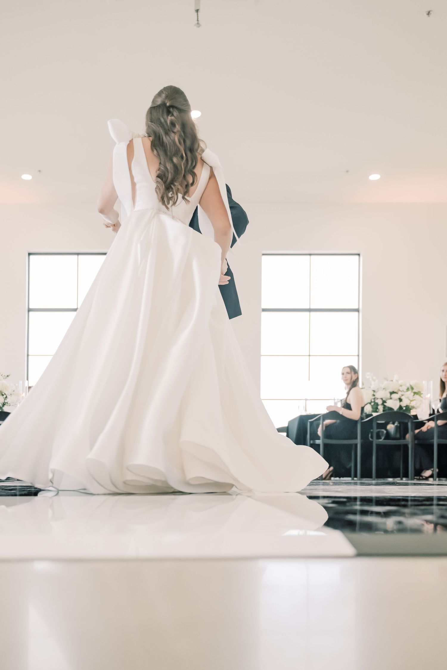 Low angle view of bride in a white ball gown with open back dancing with groom in a bright venue.
