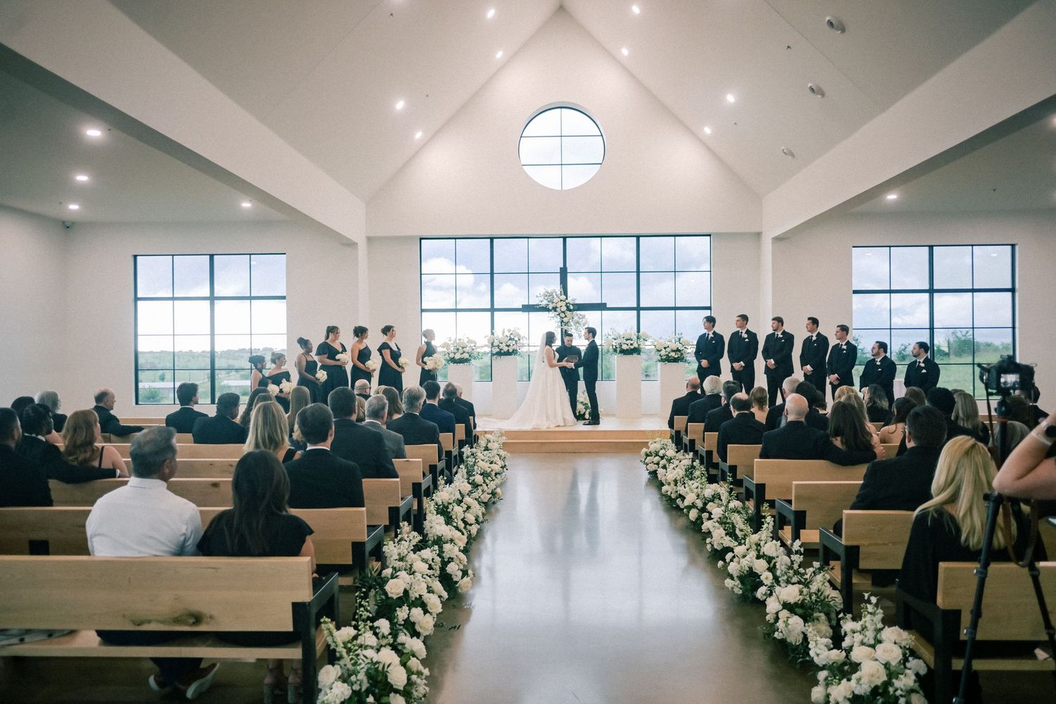 Elegant wedding ceremony in bright white chapel with wooden pews lined with white floral arrangements.
