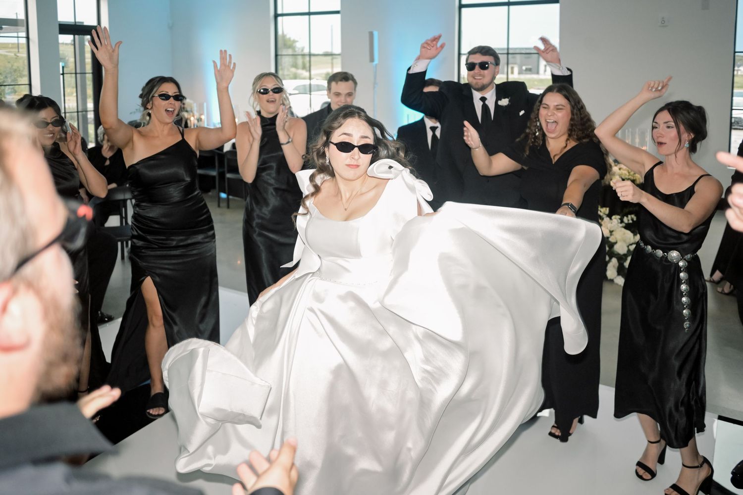 Bride in white gown dancing energetically with wedding party in black attire at reception.