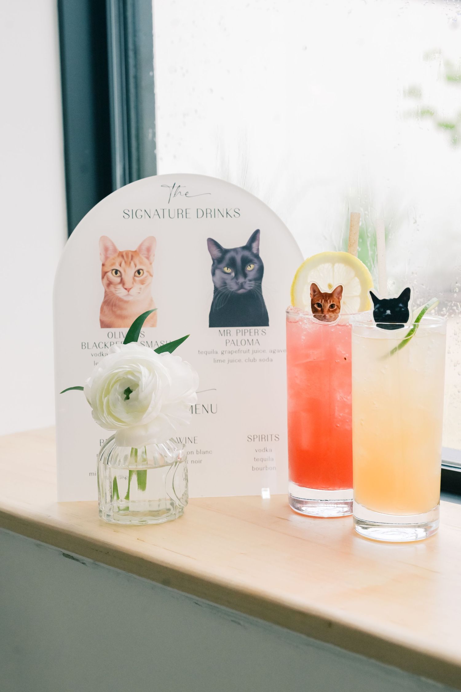 Cat-themed signature drinks menu display with two colorful cocktails garnished with cat toppers at a wedding bar.