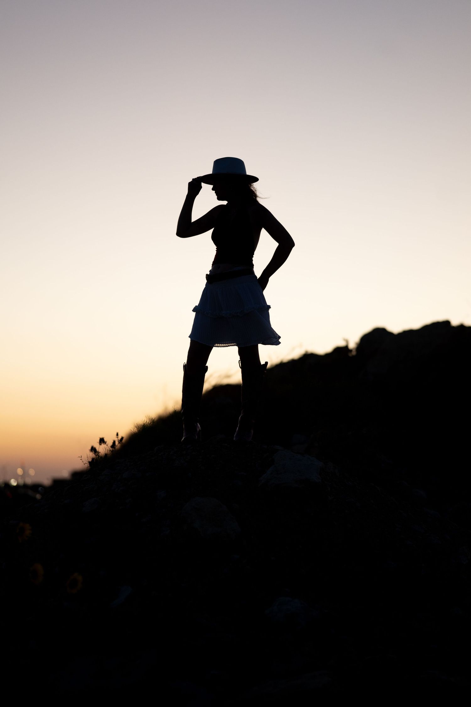 Silhouette of a woman in a skirt and hat standing on rocks against a golden sunset sky.