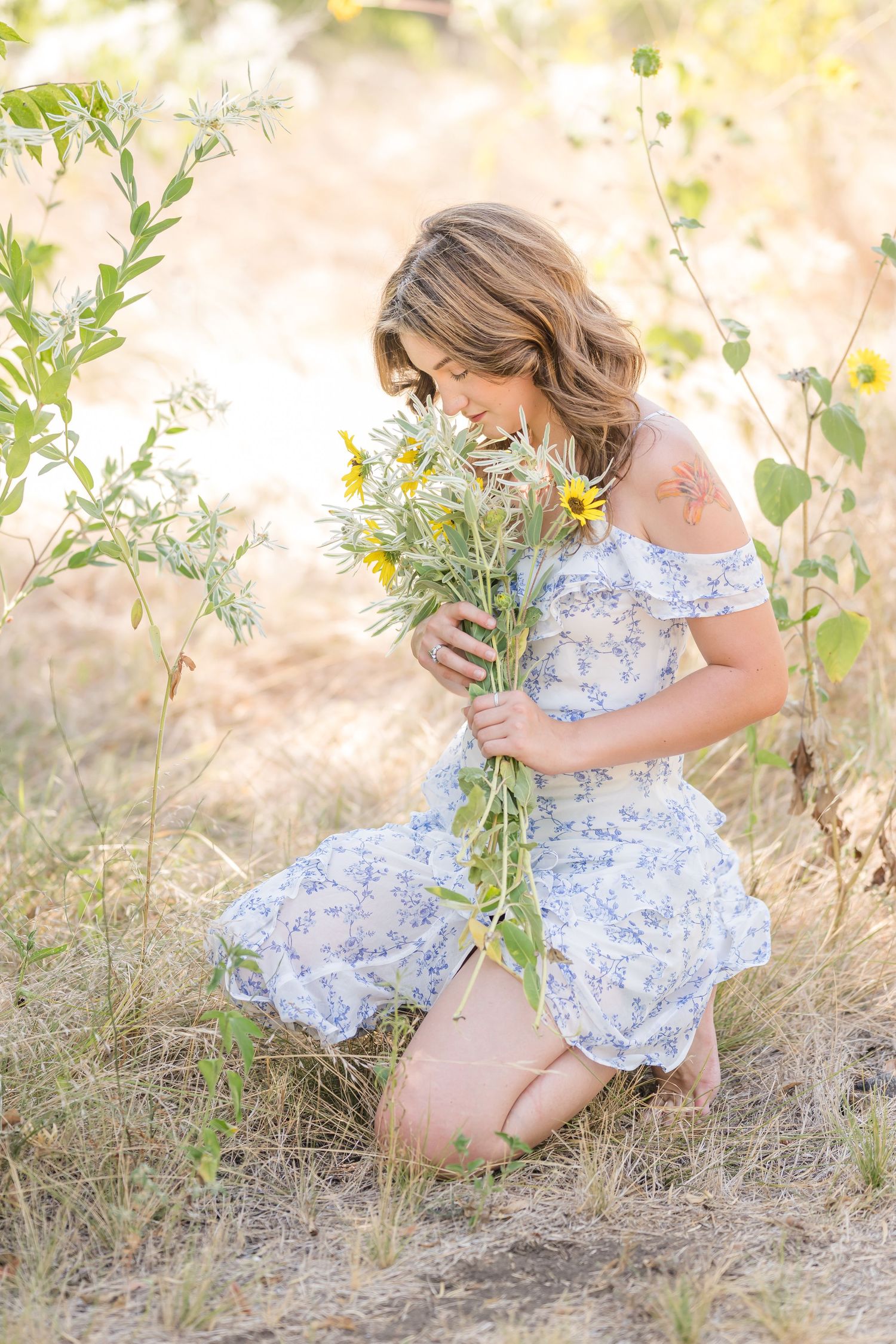 A young woman in a floral dress kneels in a sunlit field, smelling a bouquet of wild yellow flowers.