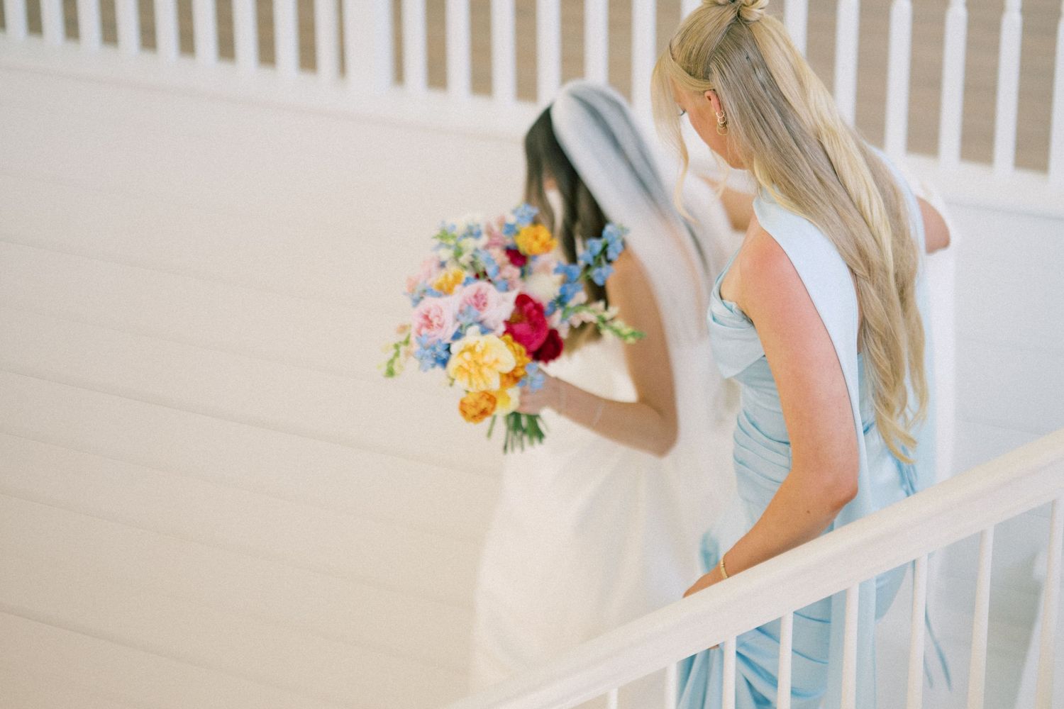 Bride holding colorful bouquet with bridesmaid in light blue dress on white staircase.