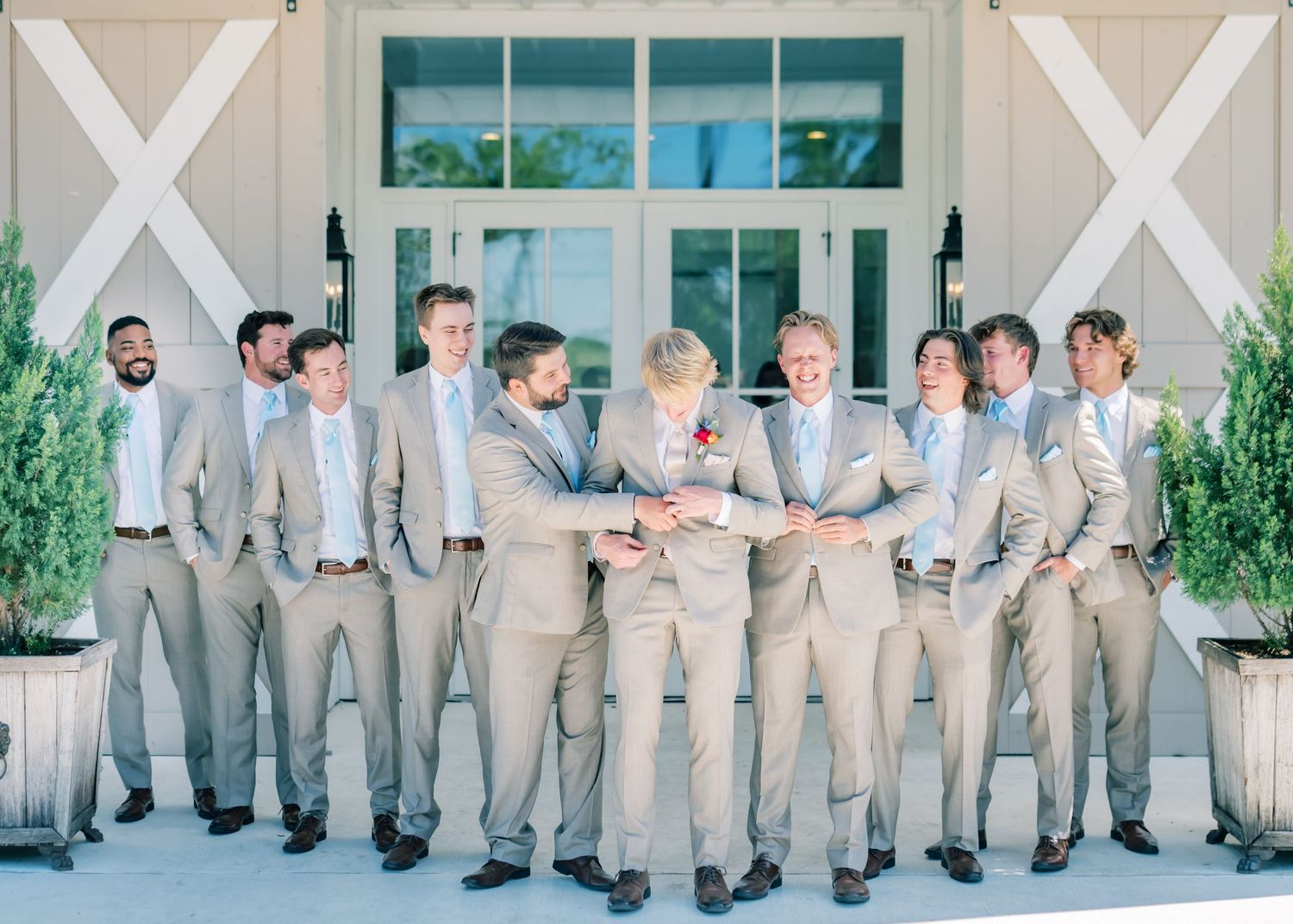 Groomsmen in tan suits pose together outside white barn doors at a wedding venue.
