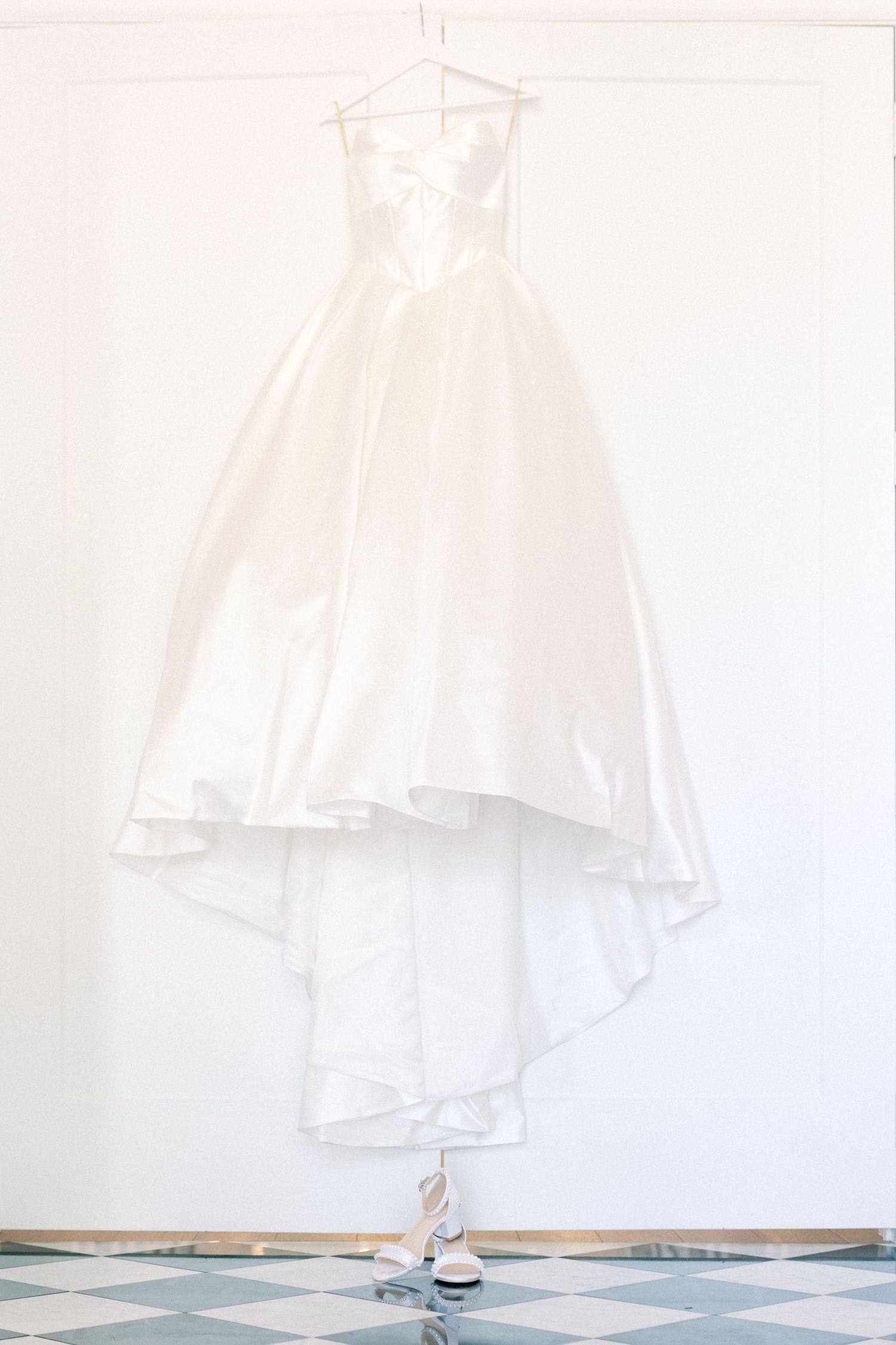 Elegant strapless white ball gown wedding dress hanging on a hanger against a white wall with bridal heels below.