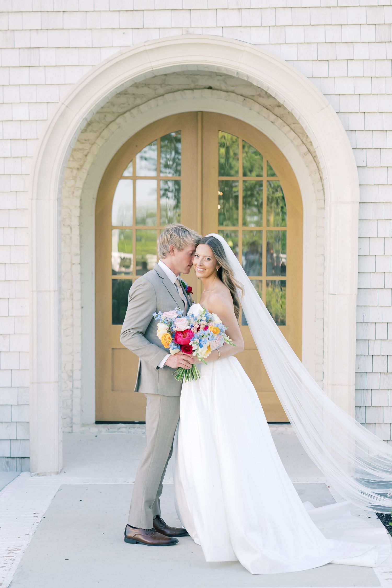 Bride and groom embrace under elegant arched doorway, her veil flowing, holding colorful bouquet at outdoor wedding venue.