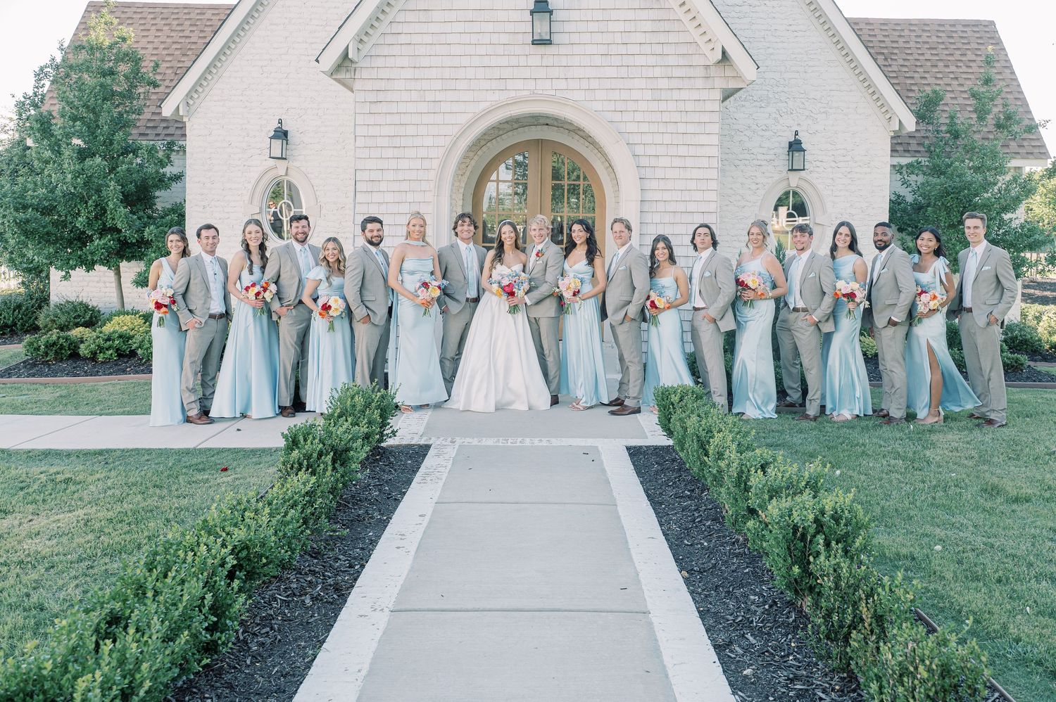 Wedding party poses outside a white stone chapel, bridesmaids in sage green gowns and groomsmen in gray suits flanking the bride and groom.