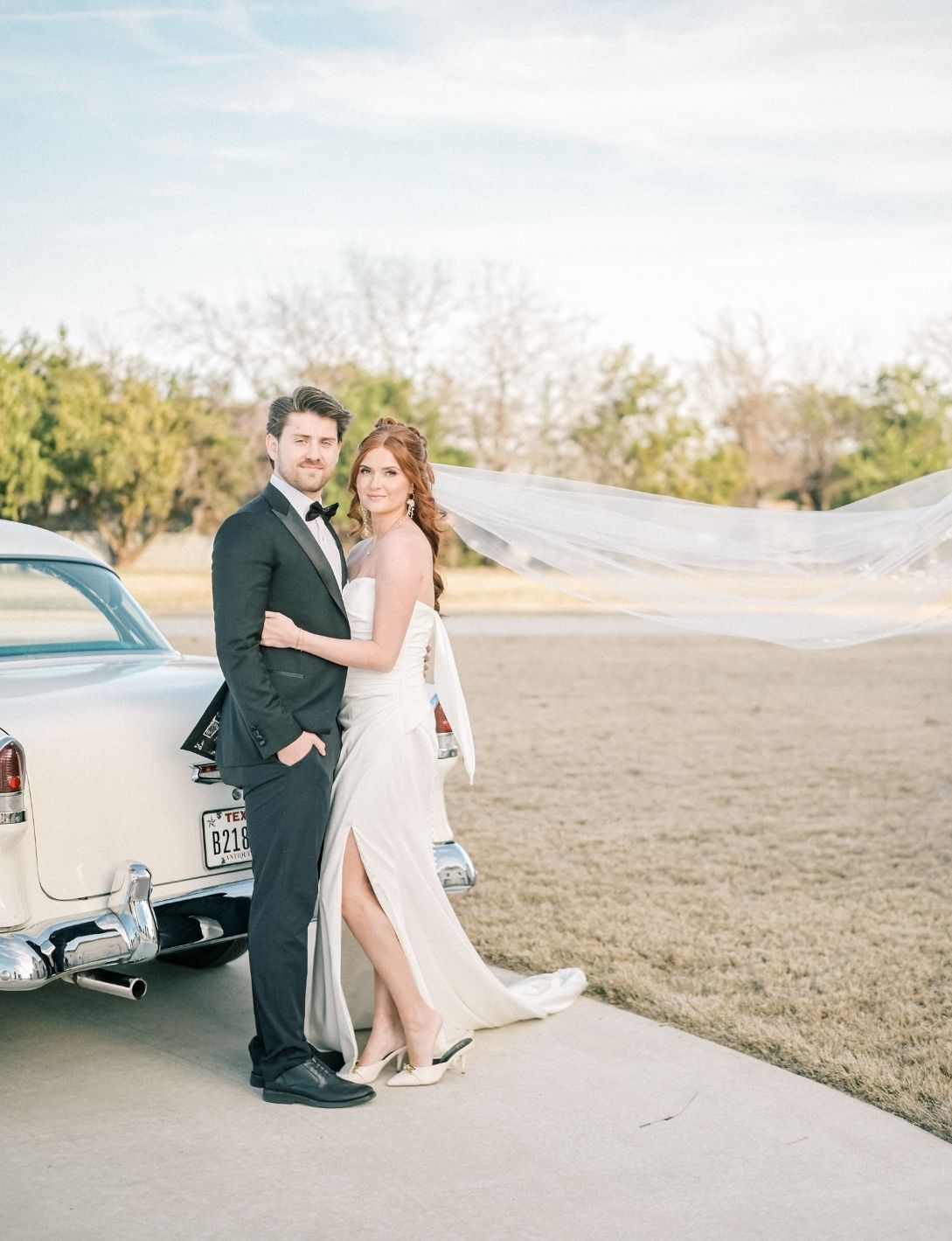 Bride and groom pose beside a vintage white car, her veil flowing in the breeze, wearing elegant wedding attire outdoors.