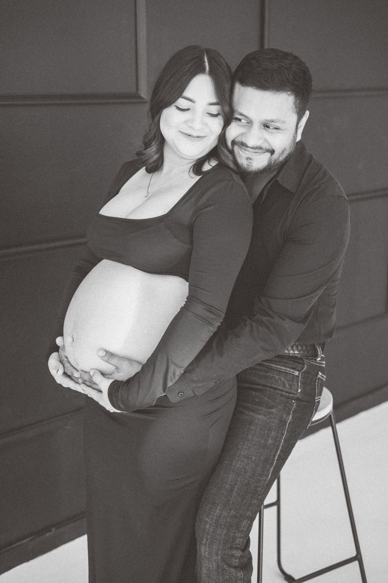 Black and white maternity photo of a smiling couple, man embracing pregnant woman from behind in a studio.