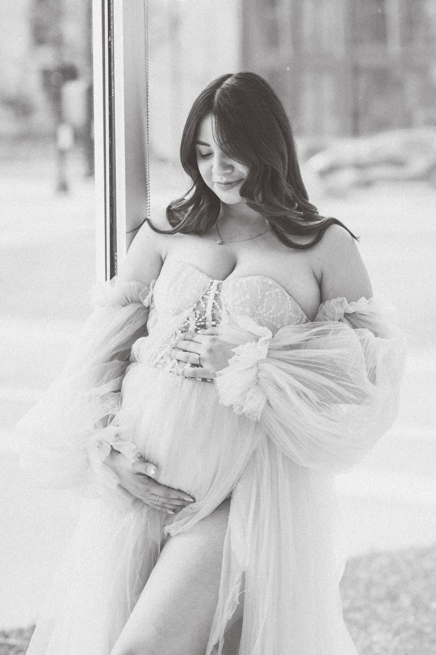 Pregnant woman in elegant off-shoulder tulle maternity gown poses gracefully in black and white photo.