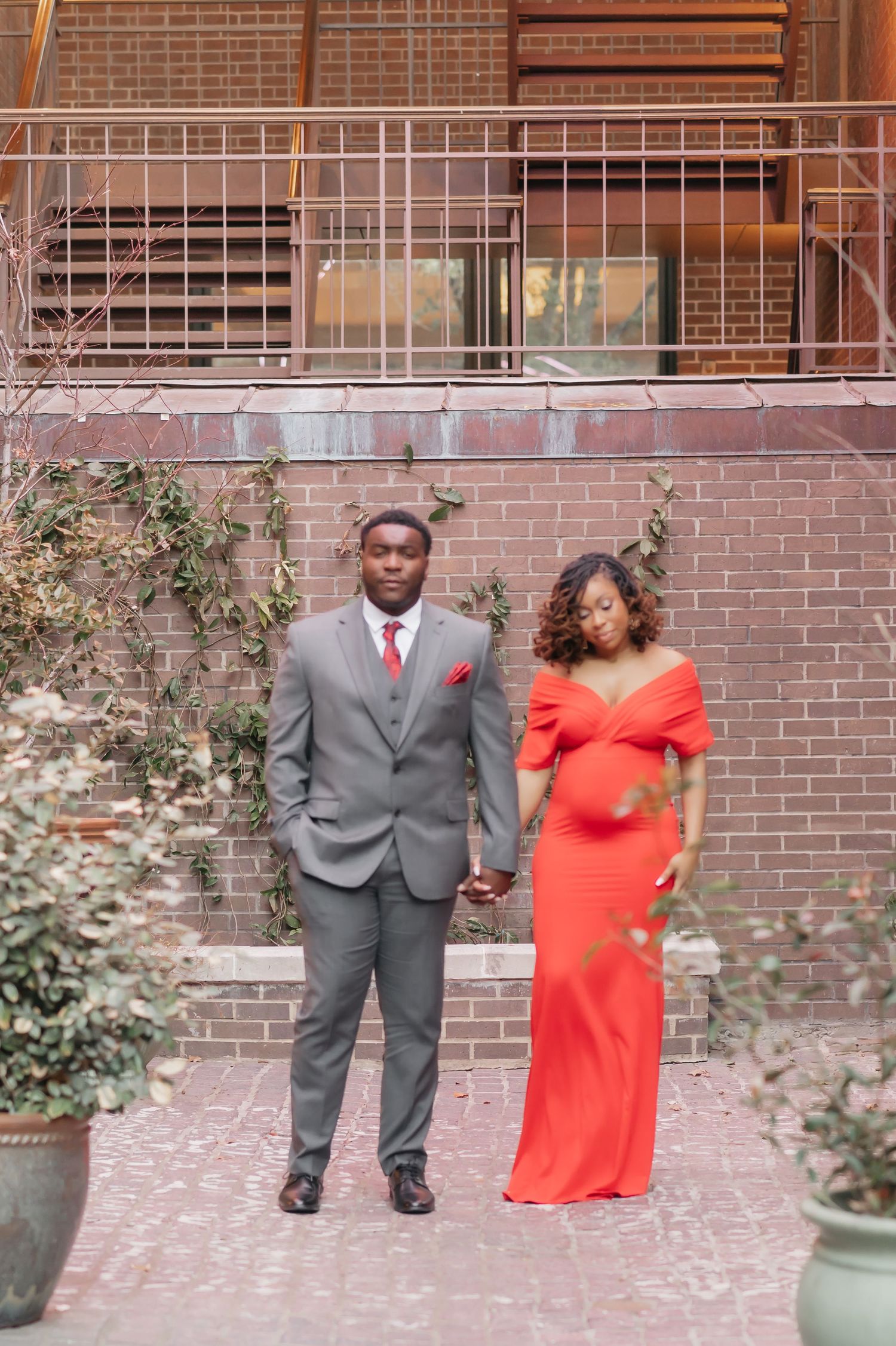 A pregnant woman in a red gown holds hands with a man in a gray suit standing before a brick wall.