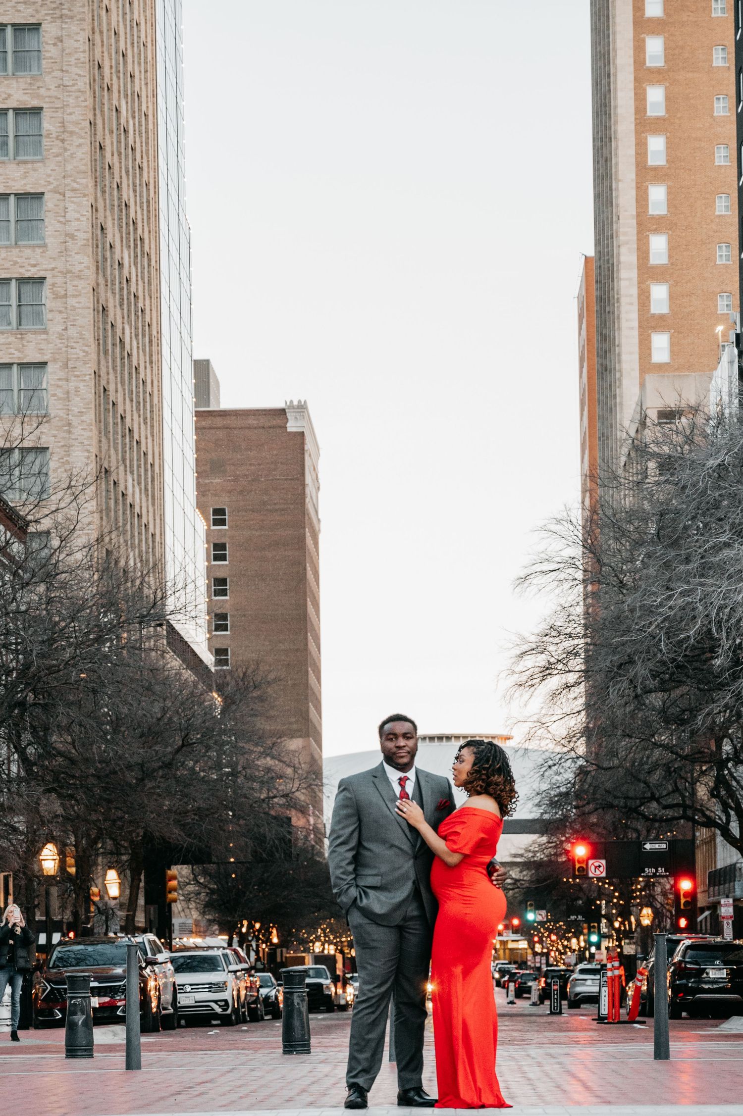 Couple posing on urban street, woman in red gown touching man's tie, city buildings backdrop.
