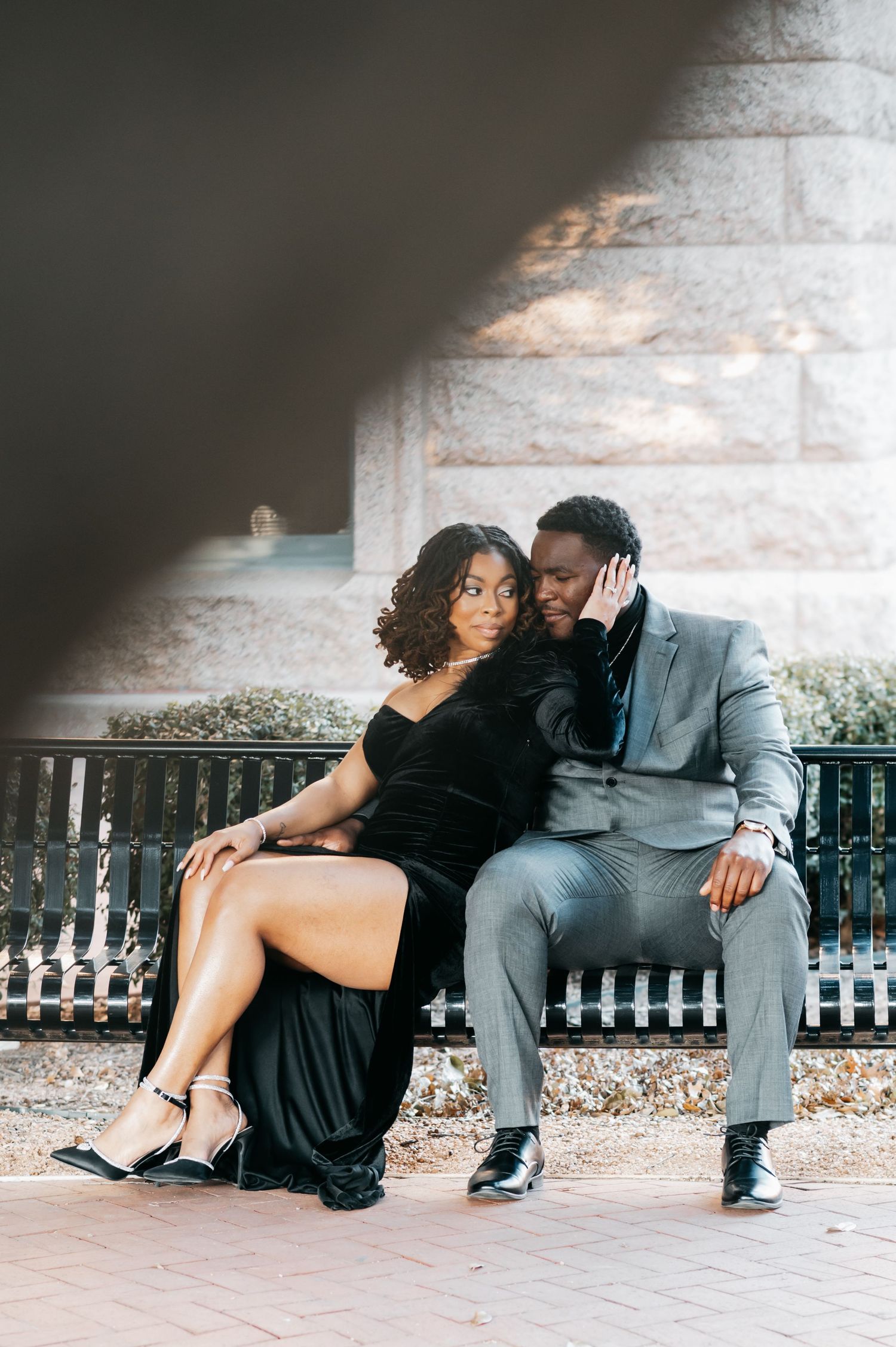 Elegant couple in formal attire sitting on a bench, woman in black gown and man in gray suit, outdoor portrait.