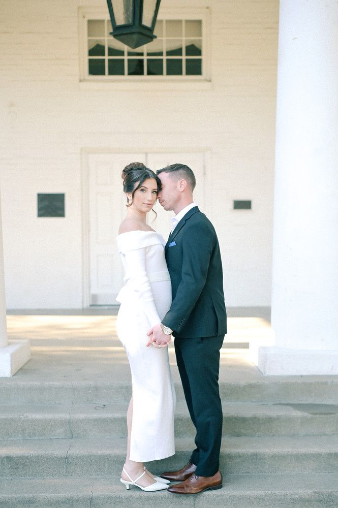 Couple in elegant wedding attire embrace on white building steps, woman in off-shoulder gown, man in dark suit.