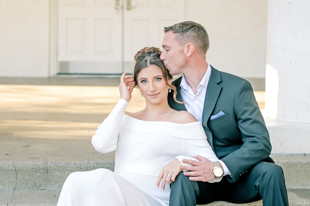 Bride in off-shoulder white gown sits with groom in gray suit on steps as he kisses her cheek tenderly.