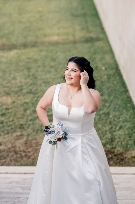 Joyful bride in white sleeveless wedding dress holds bouquet while touching her hair outdoors.
