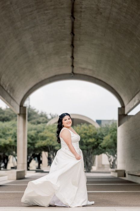 Bride in white gown twirling under concrete arch outdoors, smiling joyfully during bridal portrait session.