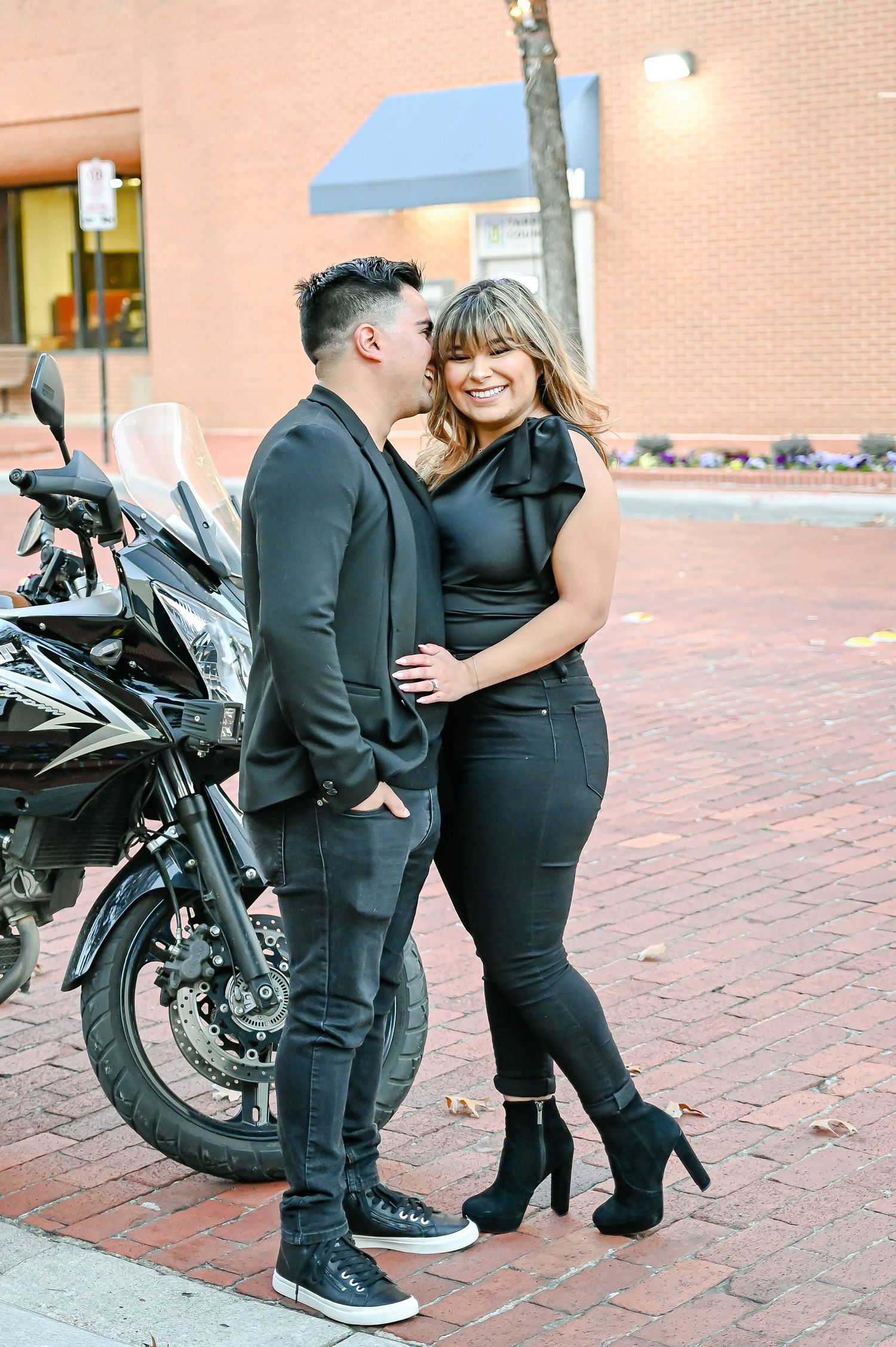 Couple in matching black outfits pose romantically beside a motorcycle on a brick sidewalk.