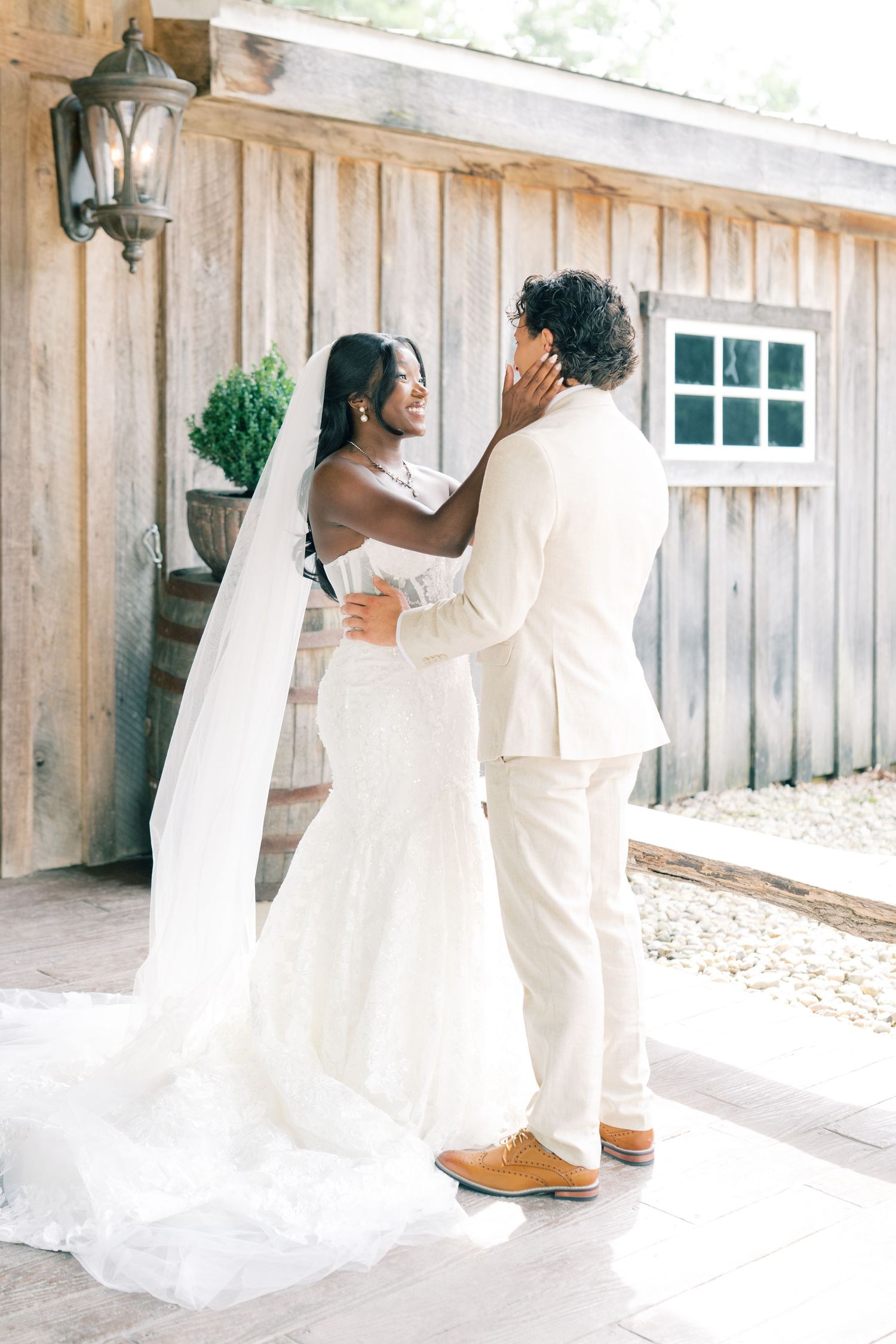 Bride and groom share a tender moment outside a rustic barn, bride wearing a white gown with long veil.