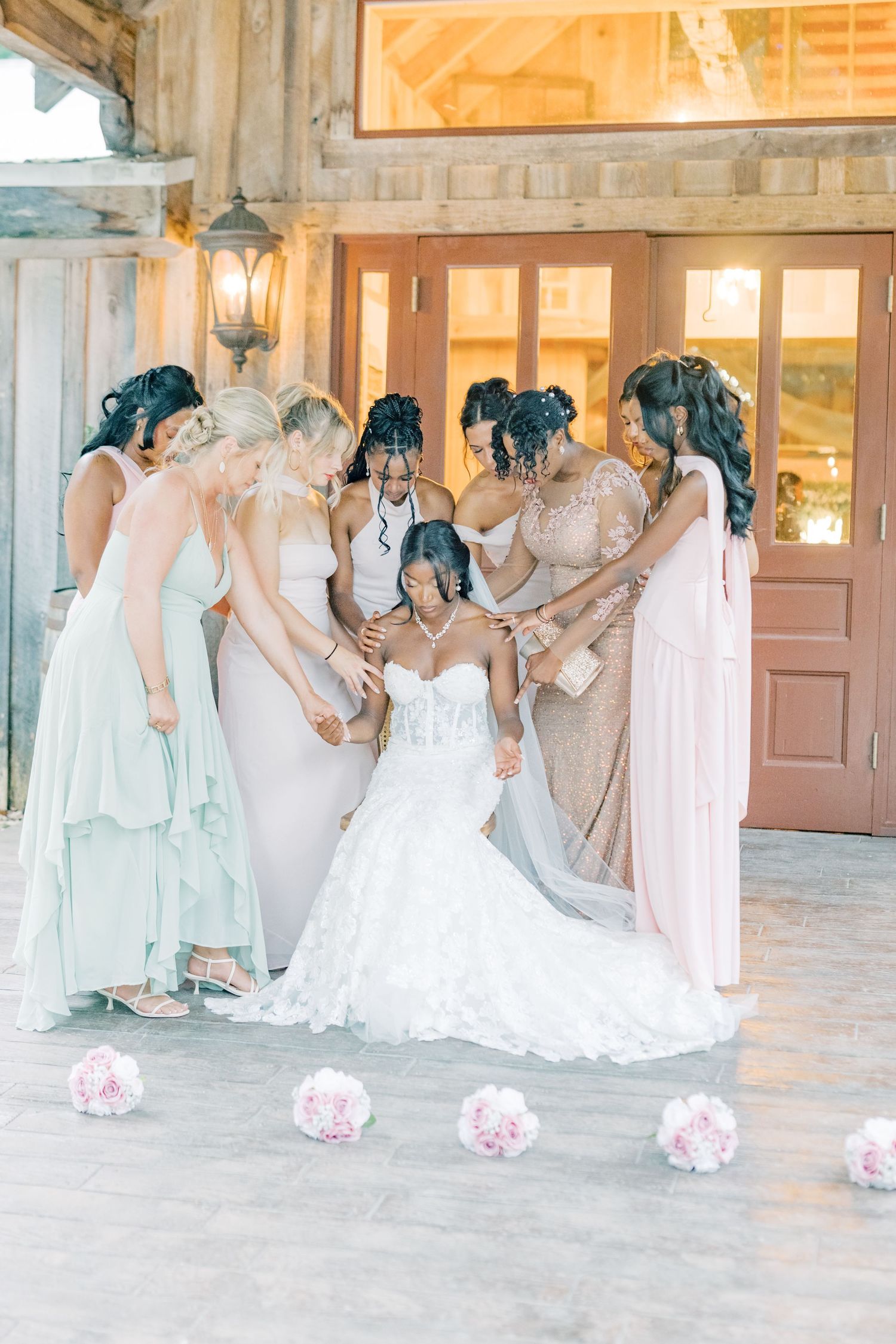Bride in white lace gown surrounded by bridesmaids in mixed pastel dresses praying together at barn venue.