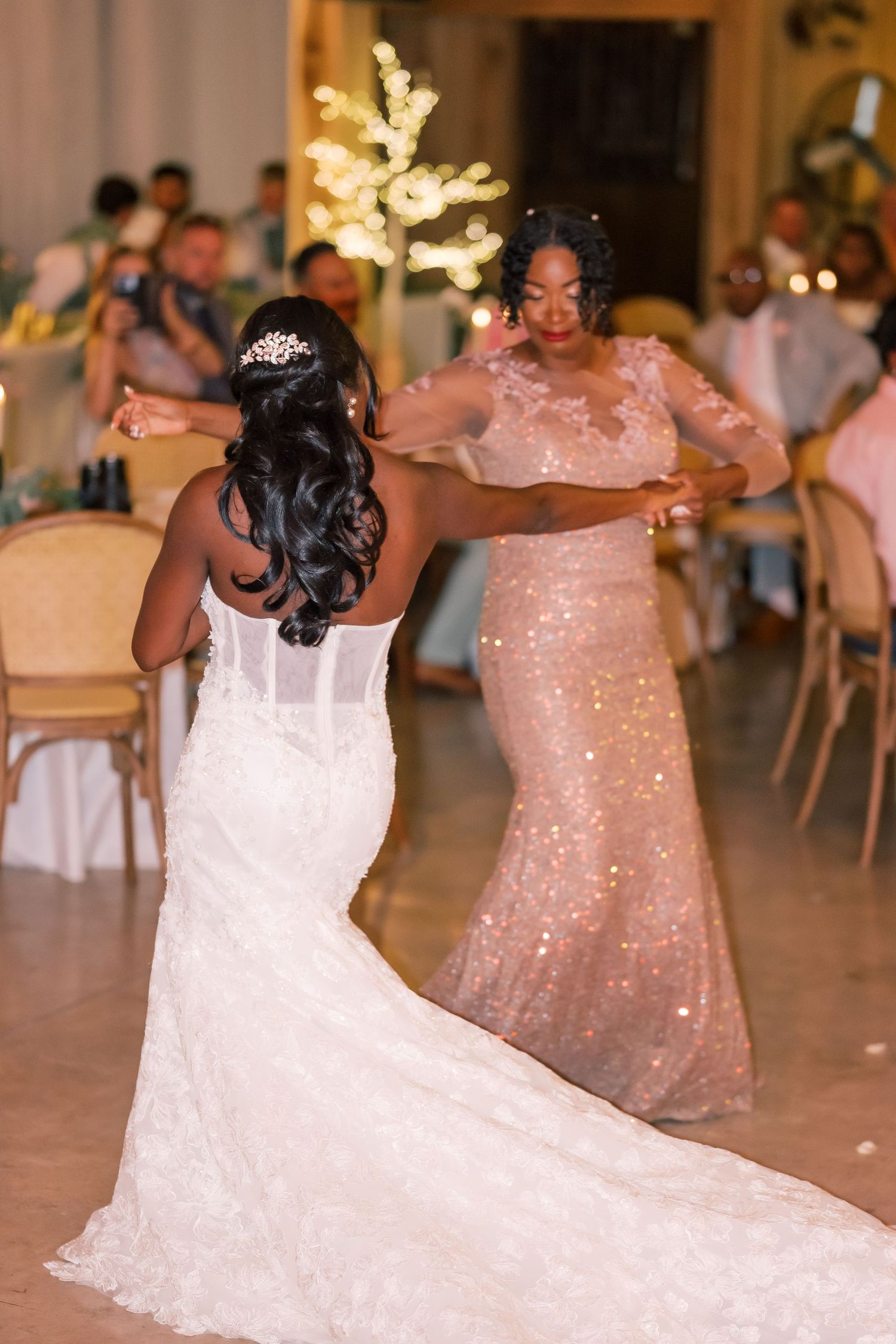 Bride in lace mermaid gown dancing with mother in sequin dress during wedding reception with warm fairy lights.