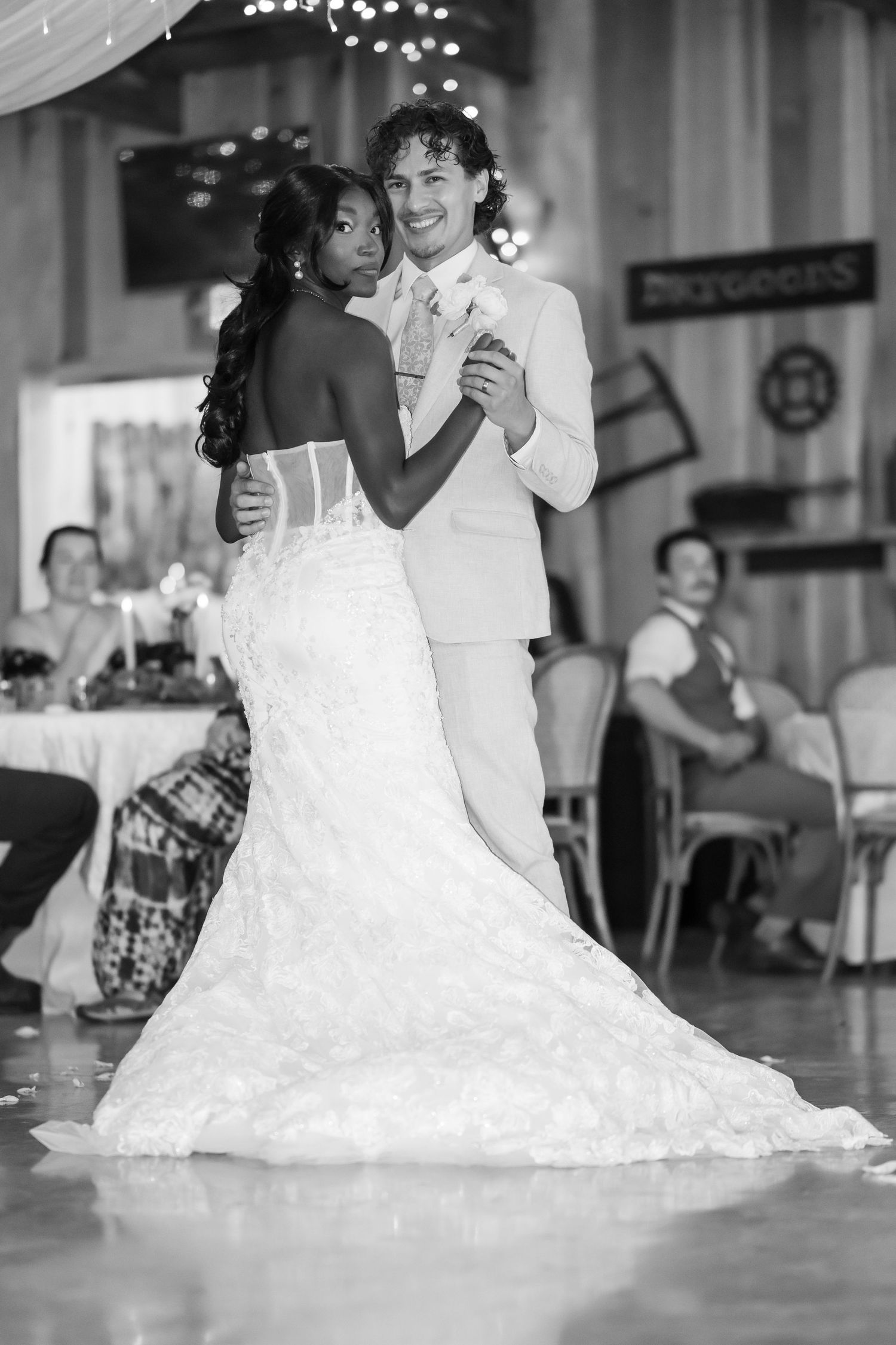 Bride and groom share their first dance in black and white photo, guests watching in rustic barn reception.