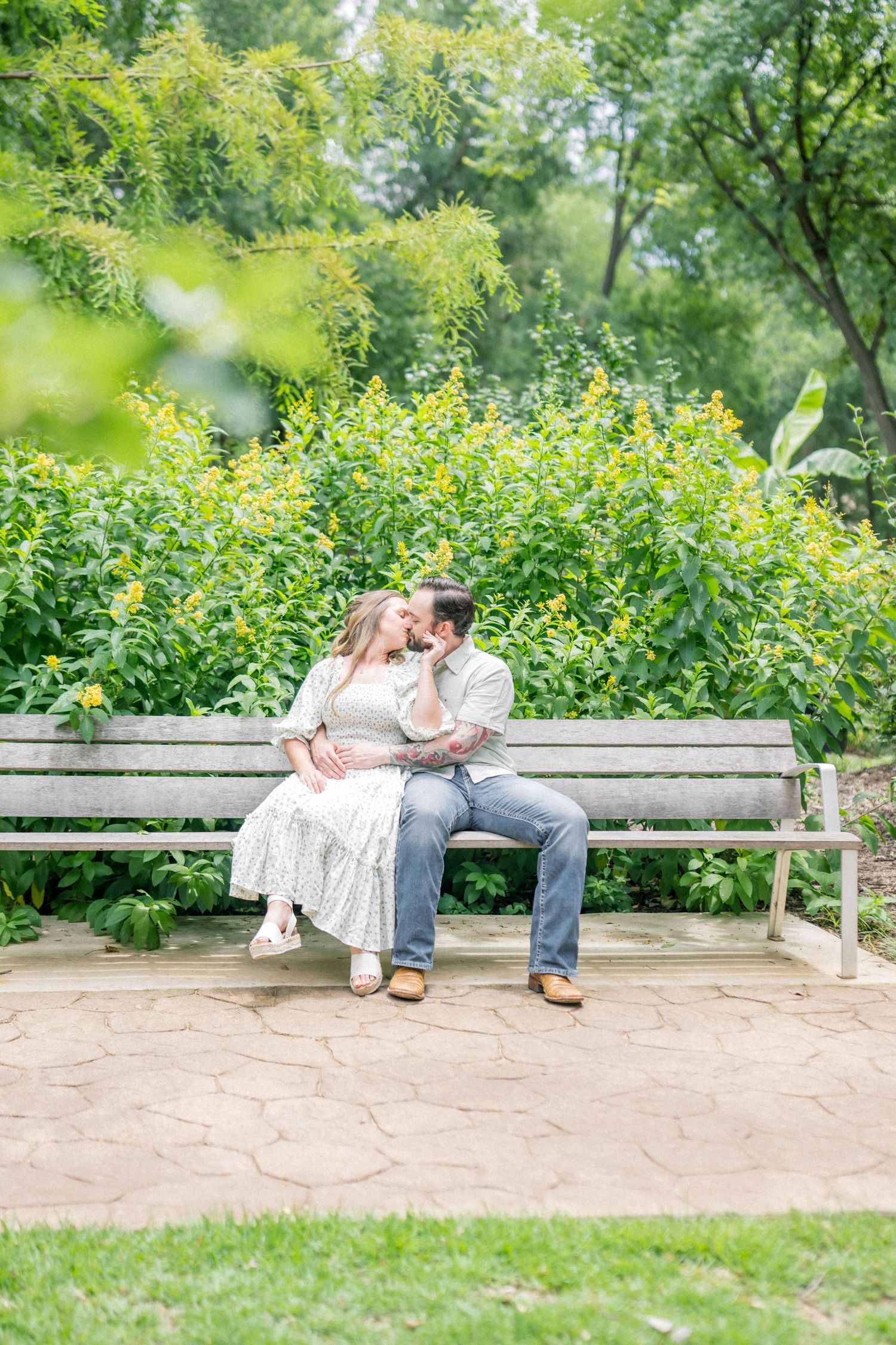 Couple shares a romantic kiss on a wooden bench surrounded by lush yellow flowering garden bushes.
