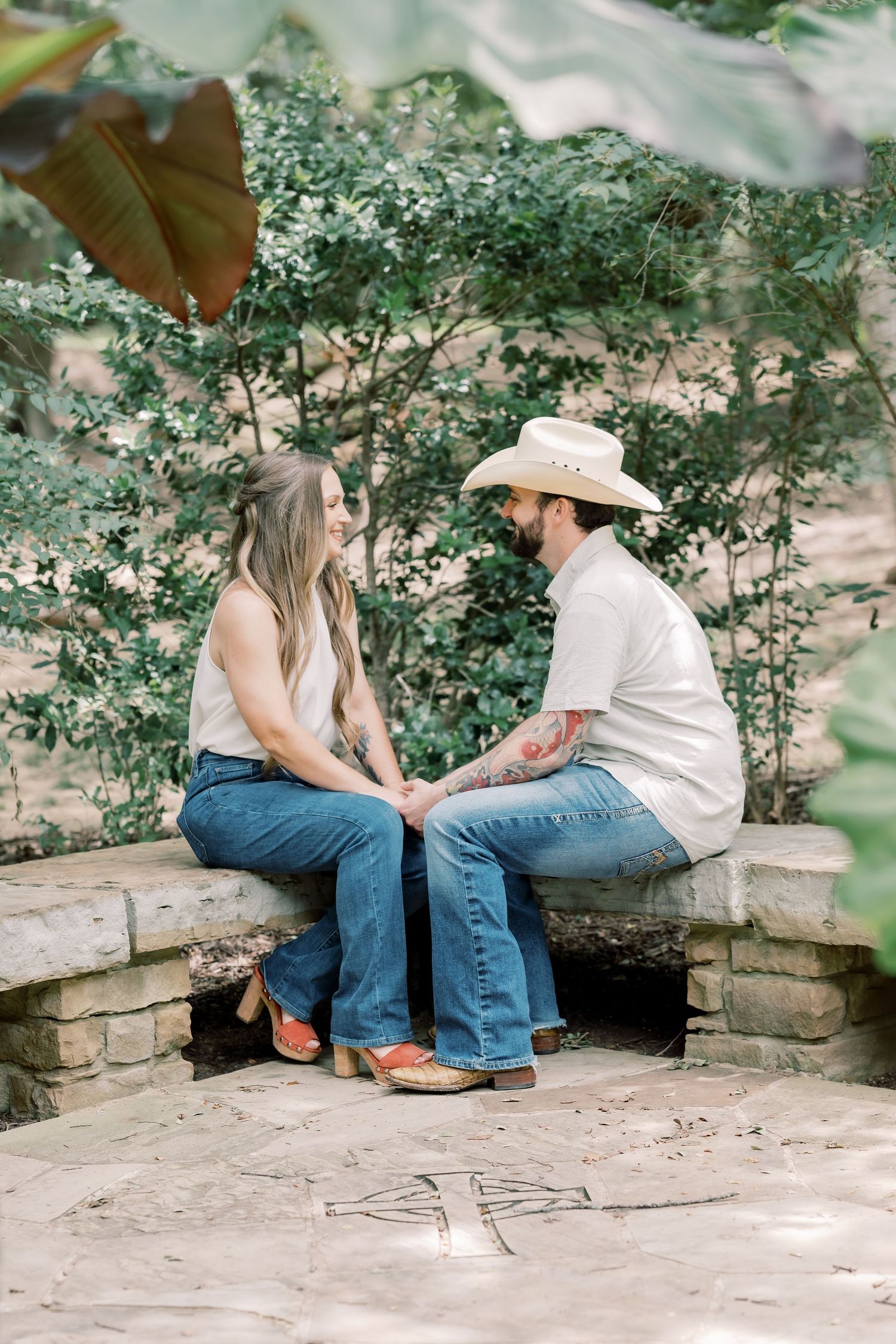Couple in cowboy hats and jeans holding hands on a stone bench in a lush garden.