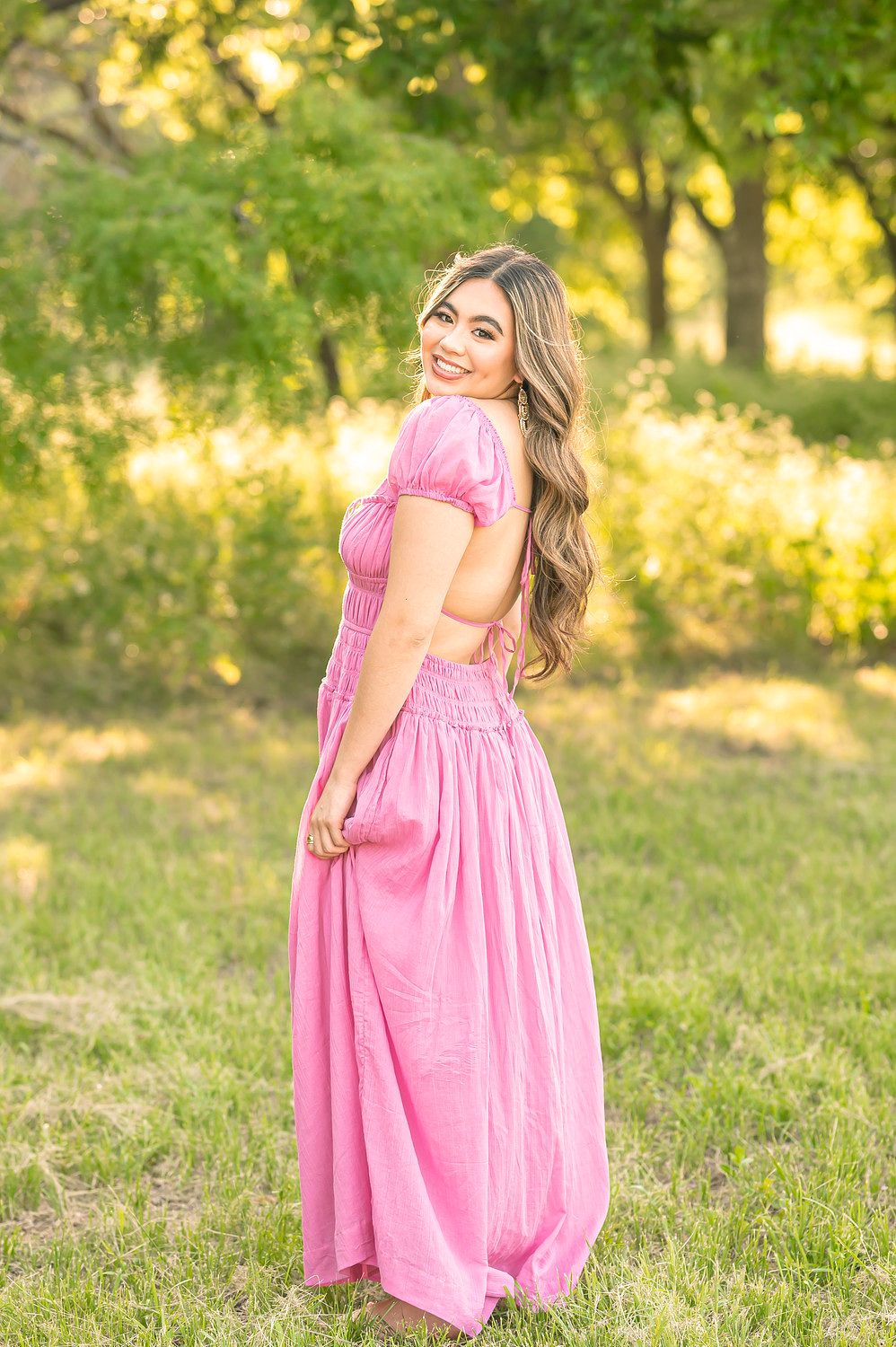 Young woman in a pink maxi dress smiling over her shoulder outdoors in a sunlit green park setting.