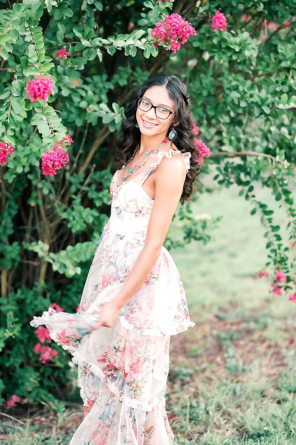 Smiling young woman in floral dress and glasses posing under pink bougainvillea arch in lush green garden. Seymour TX HS Senior Photography