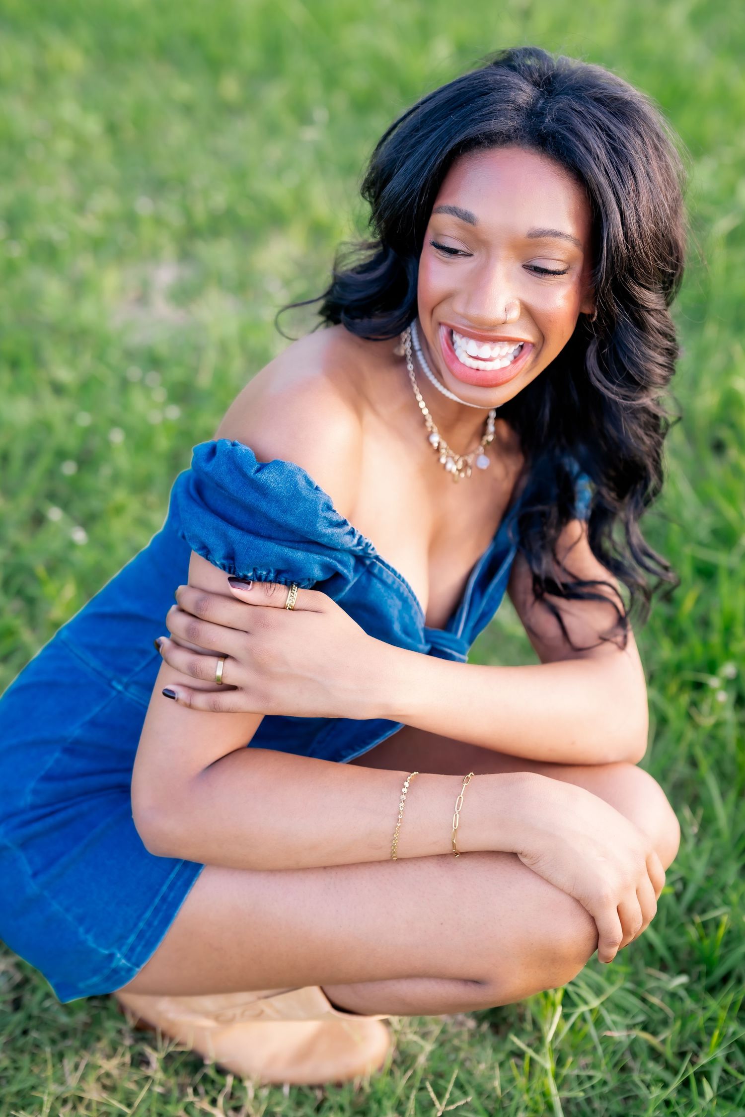 Young woman in blue off-shoulder dress crouching on grass, laughing joyfully with jewelry accessories.