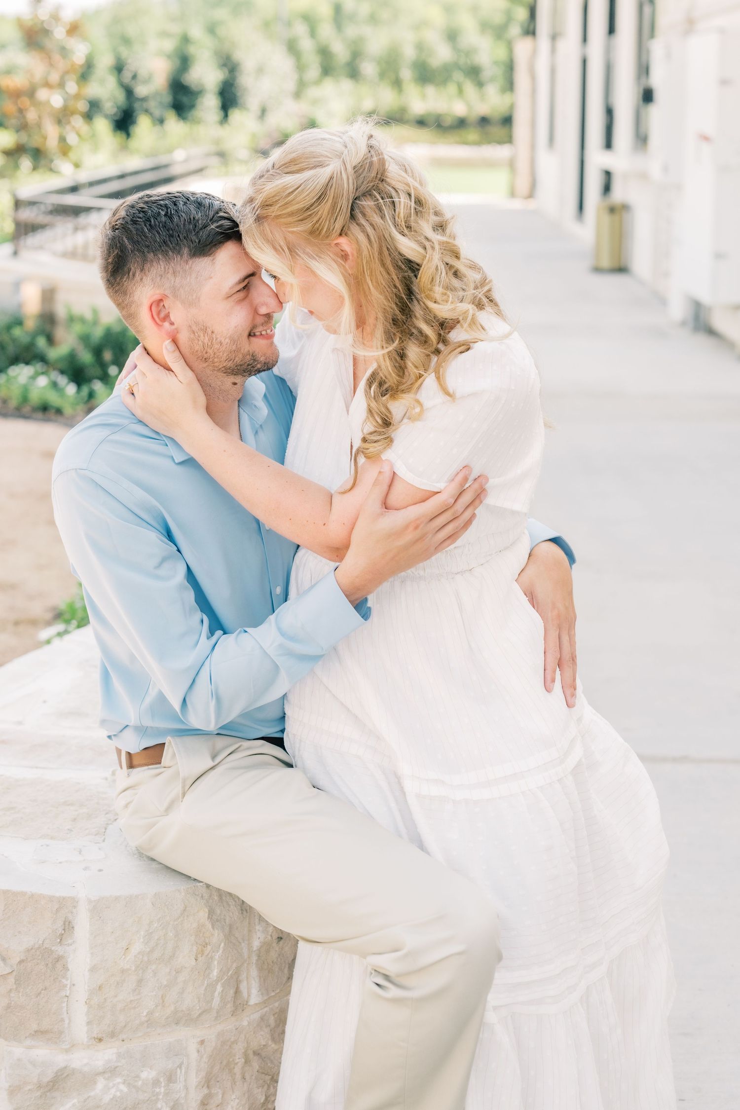 Romantic couple embracing and touching foreheads while sitting on a stone wall outdoors at a scenic venue.