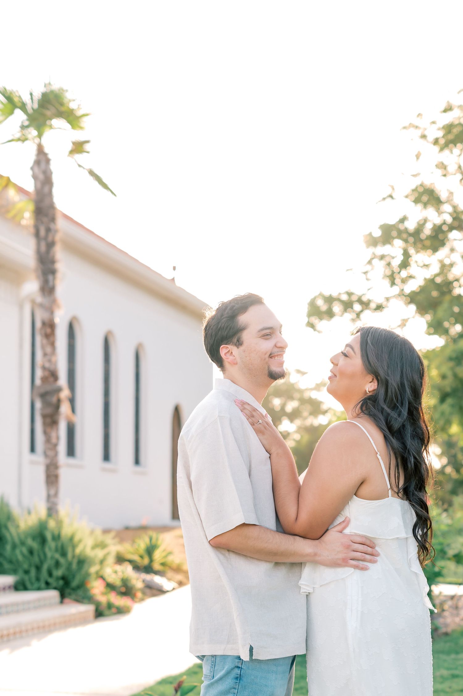 A couple shares a joyful moment laughing together outdoors near a white building with palm trees at sunset.
