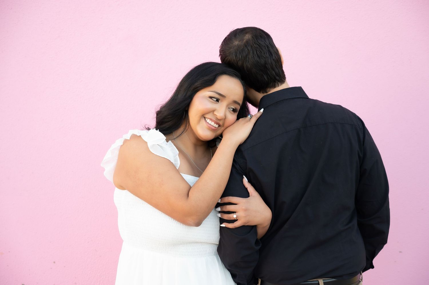 Romantic couple embracing against pink wall with woman in white dress and man in dark shirt.