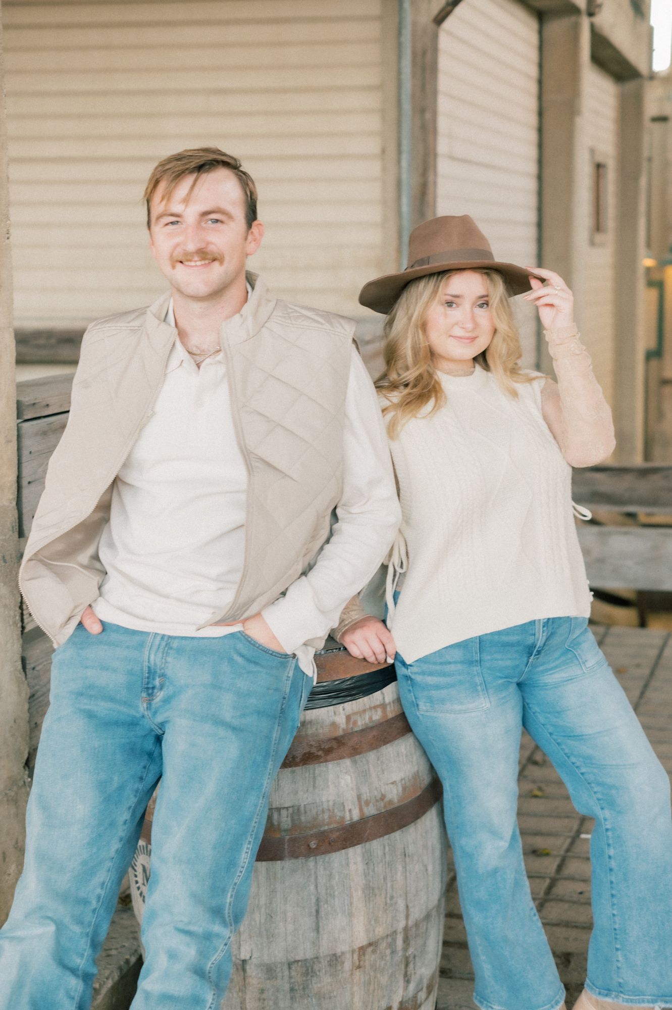 A couple in casual western attire share joyful moments leaning against wooden barrels in multiple poses.