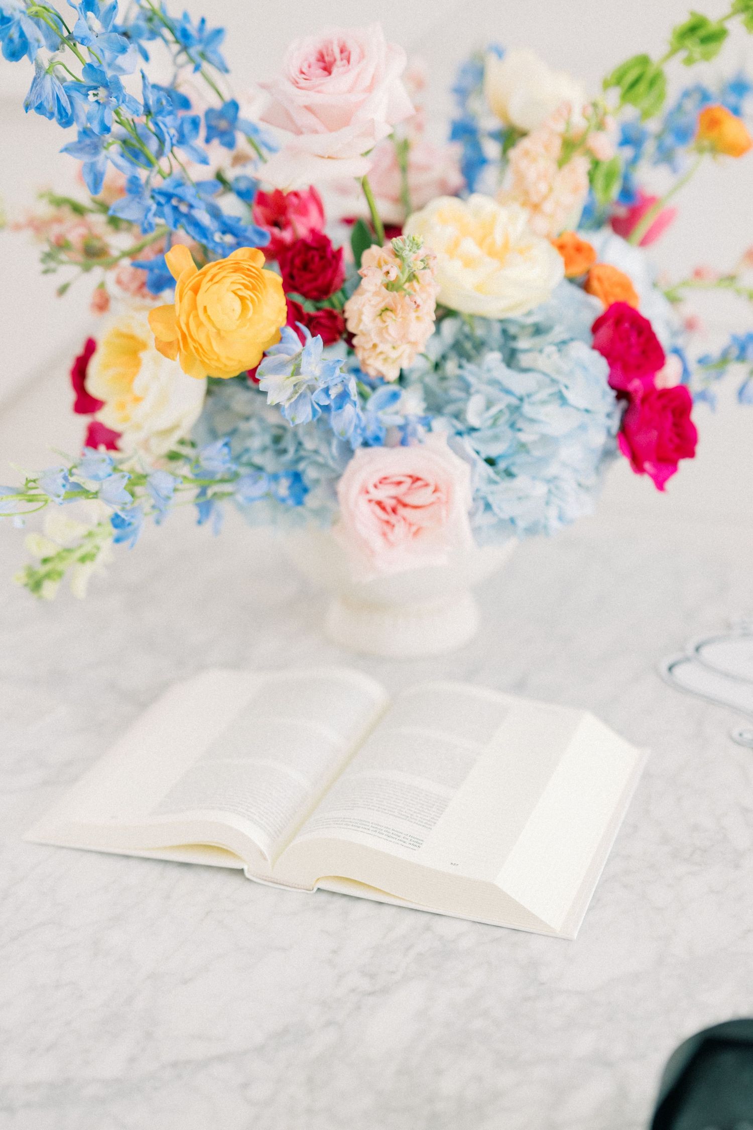 Open book on marble table beside colorful floral arrangement with pink roses, blue hydrangeas, and yellow ranunculus.