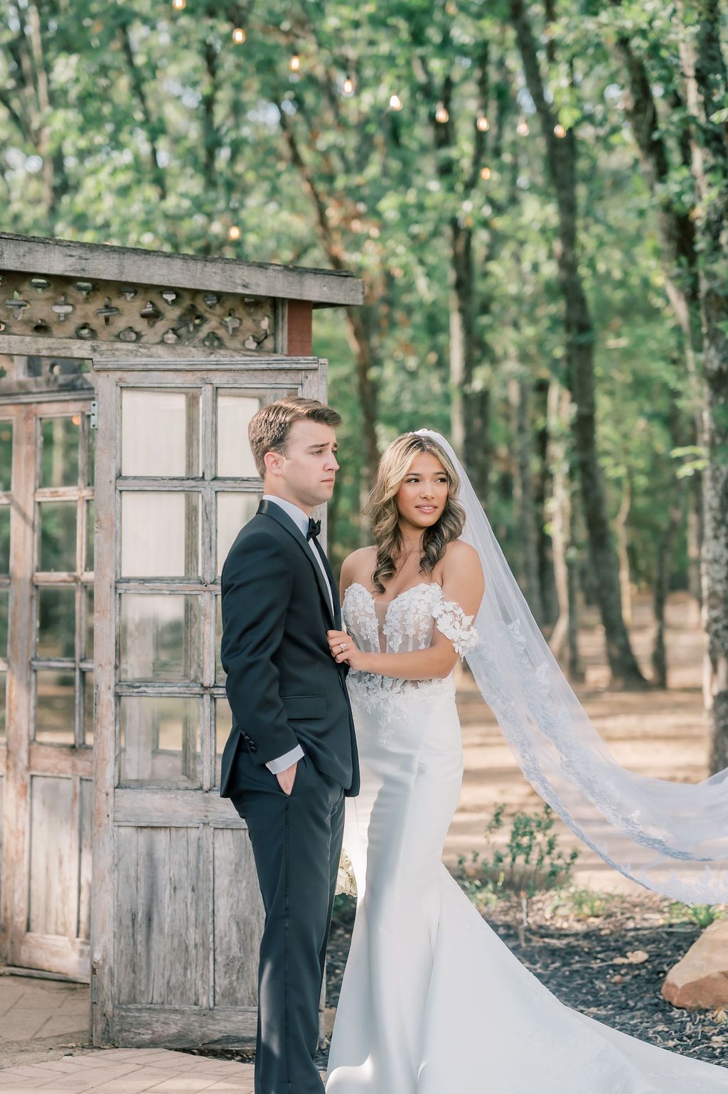 Bride in lace gown with flowing veil stands with groom in black tuxedo by rustic wooden doors outdoors.