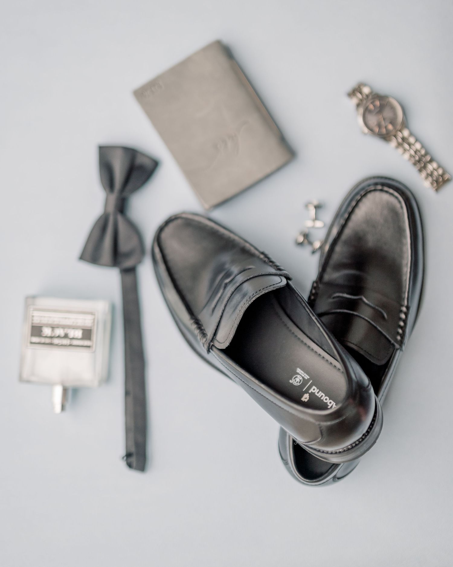 Groom wedding day accessories flat lay with black loafers, bow tie, watch, cufflinks, and gray wallet.