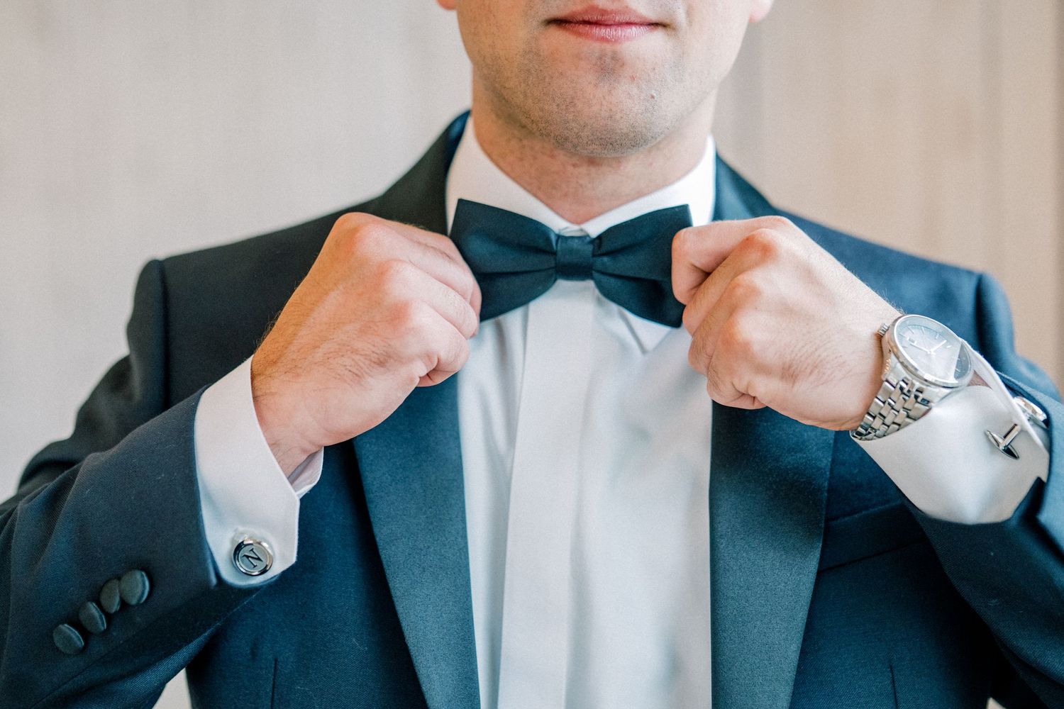 Groom in navy tuxedo adjusting his black bow tie, wearing a silver watch and cufflinks.