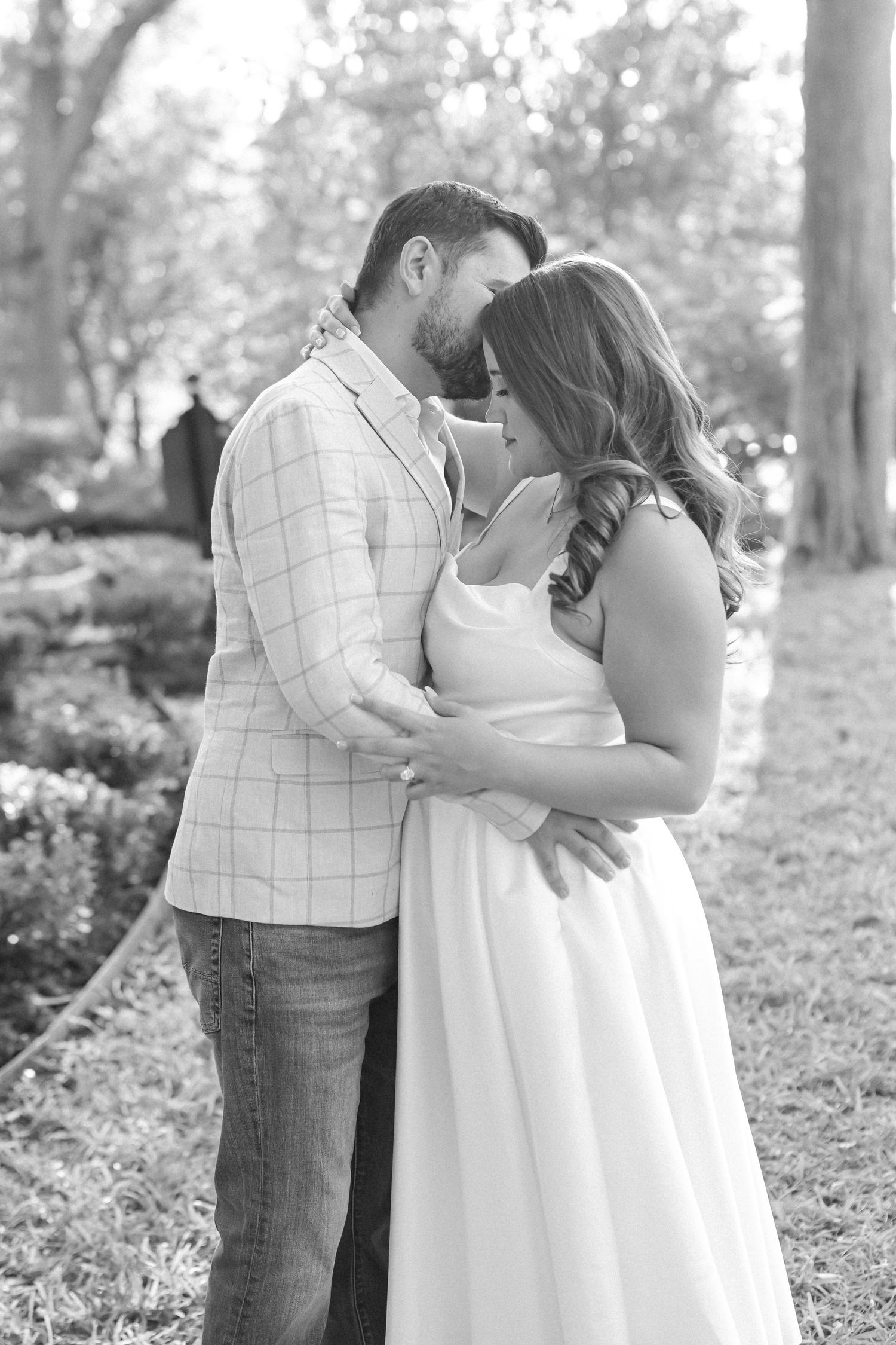 Black and white engagement portrait of a man kissing his partner's forehead as they embrace in a park with tall trees.