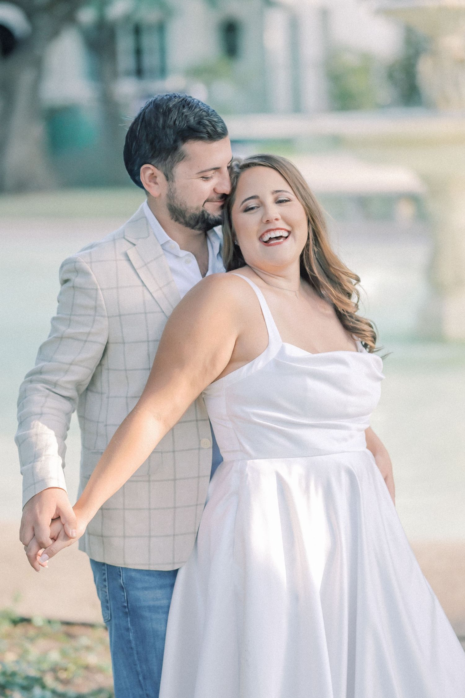 Smiling woman in a white dress holds hands with a man in a plaid blazer as he nuzzles her cheek during an outdoor engagement photo session.