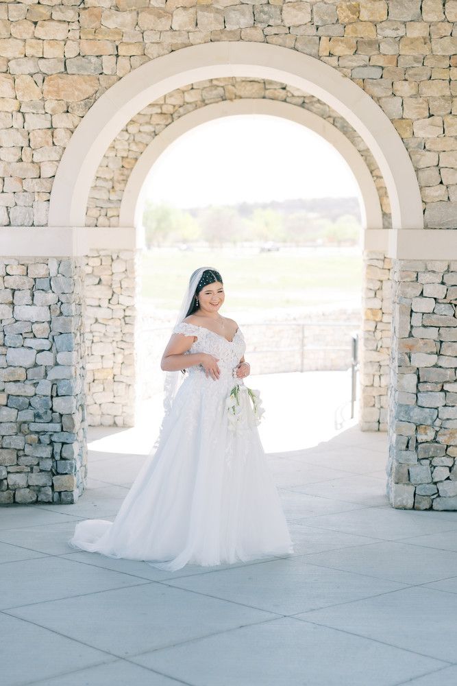 A bride in a classic white dress poses beneath a series of stone arches with natural light streaming in.