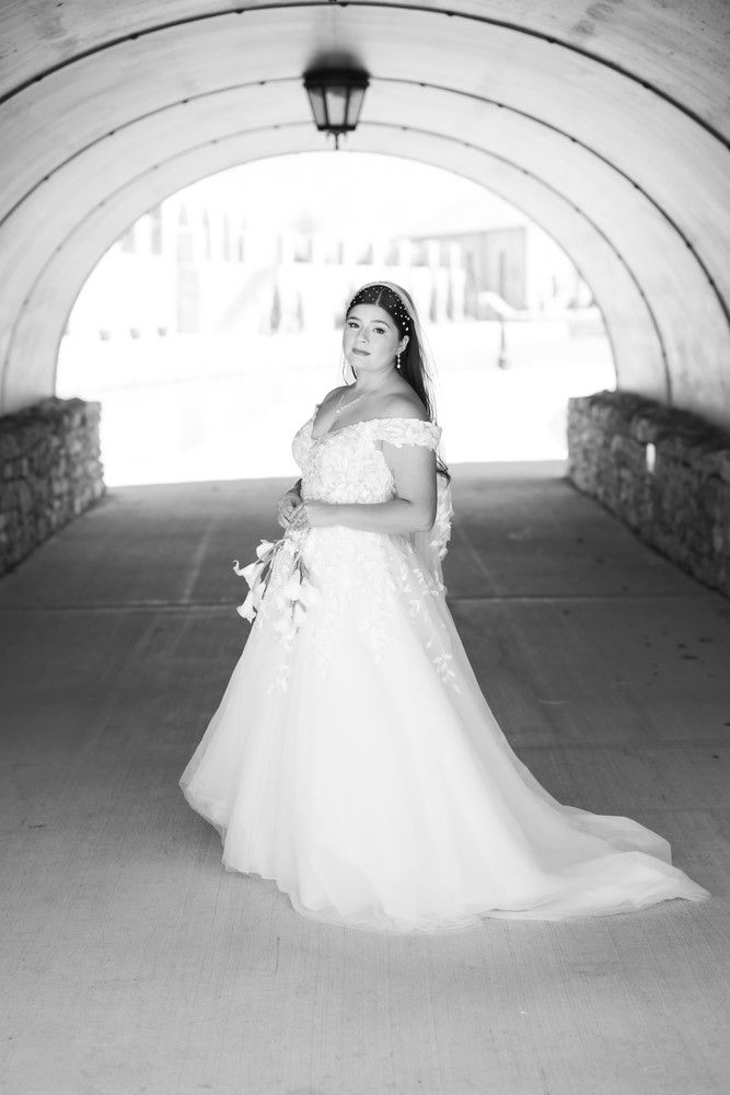 A bride in an off-shoulder white wedding gown stands elegantly in a stone archway tunnel, captured in black and white.