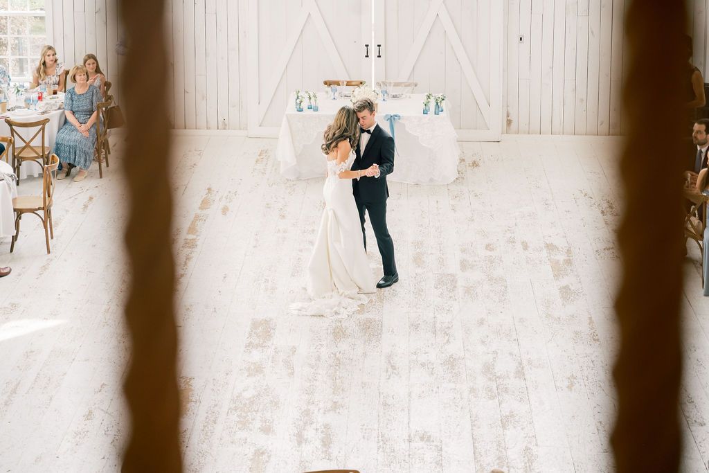 Bride and groom share first dance on white wooden floor inside bright airy barn wedding venue with guests seated nearby.