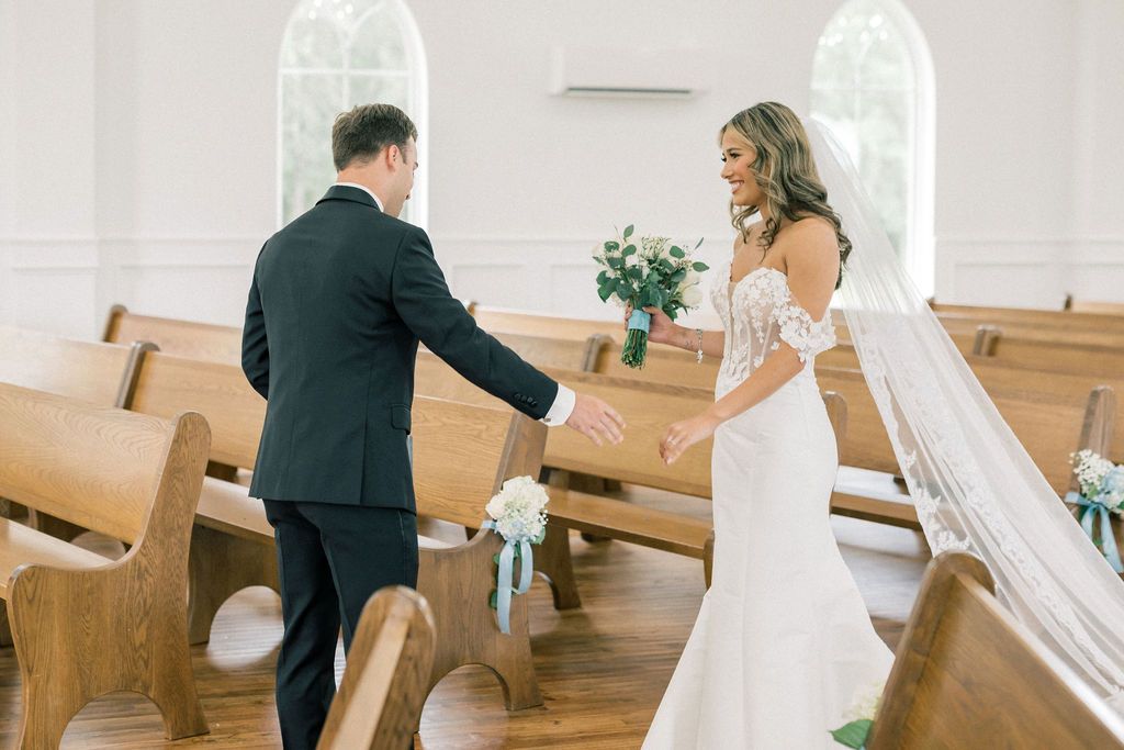 Bride and groom share a tender first look moment inside a white chapel with wooden pews on their wedding day.