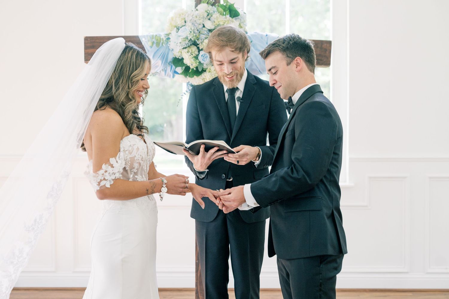 Bride and groom exchange rings during intimate wedding ceremony officiated by minister at floral altar.